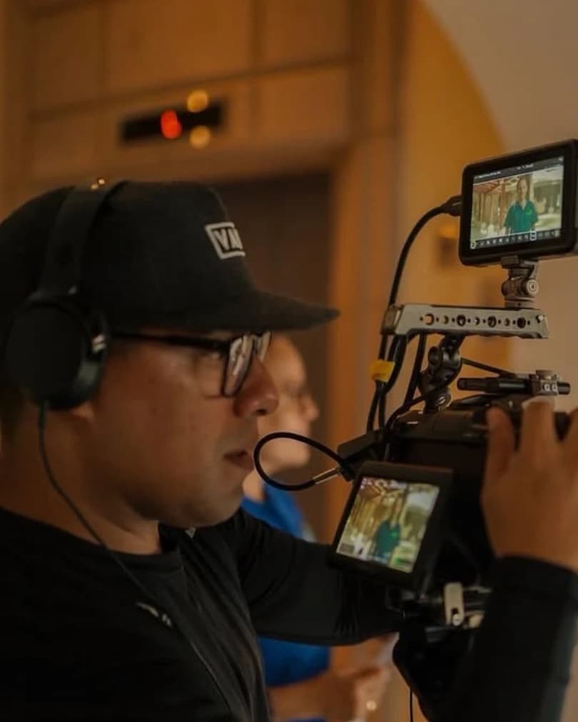 Camera operator in a cap and headphones running a cinema camera with an external monitor during an indoor shoot